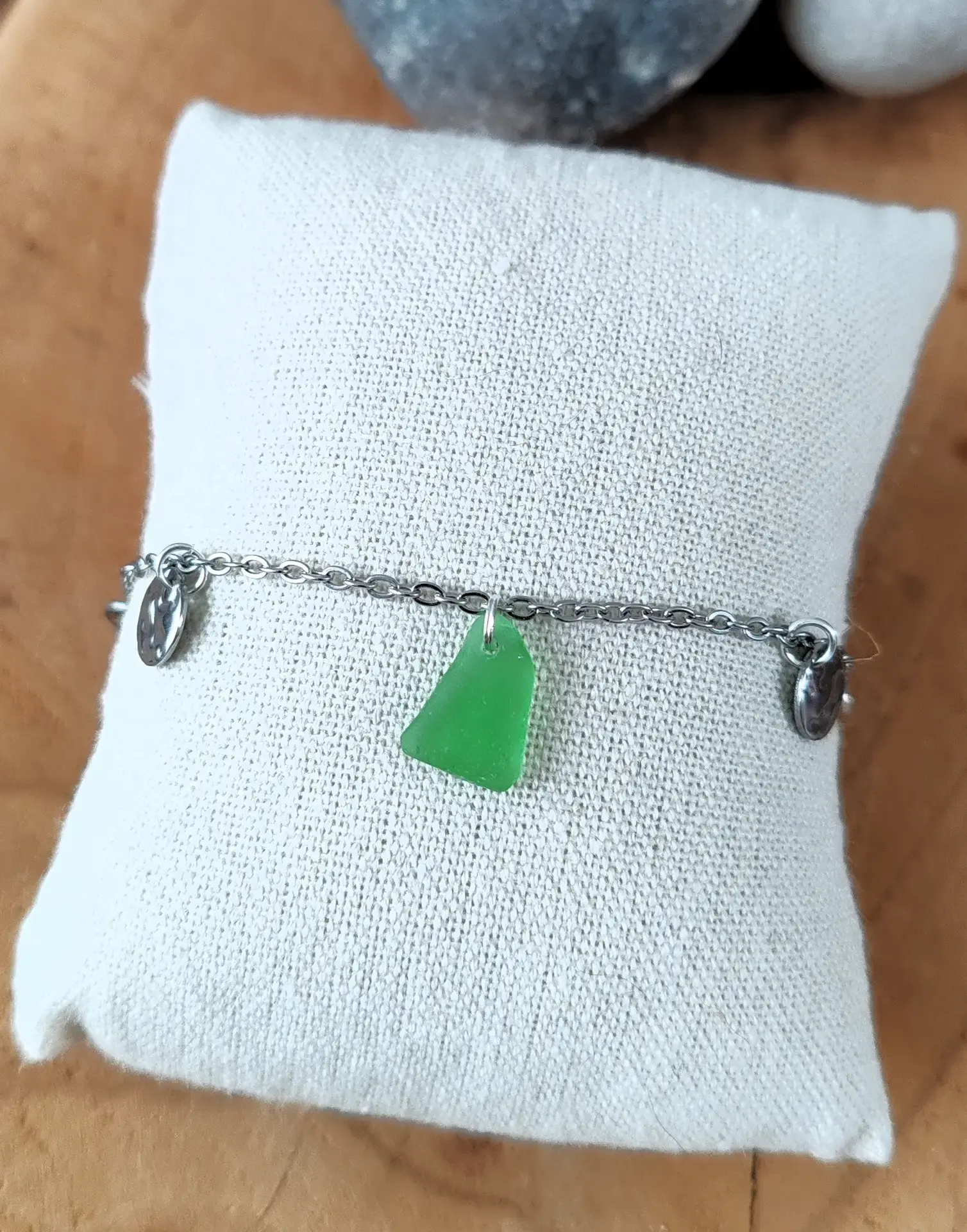 Le bracelet Pelagos argenté et son éclat de verre de mer vert, posé sur un coussin de présentation en lin clair